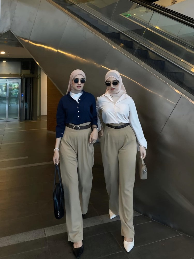 Foto de duas mulheres ao lado de escada rolante. A da esquerda usa blusa azul de manga longa, calça pantalona bege e hijab bege. A da direita também usa pantalona e hijab bege, mas blusa de manga longa branca. Ambas usam óculos de sol