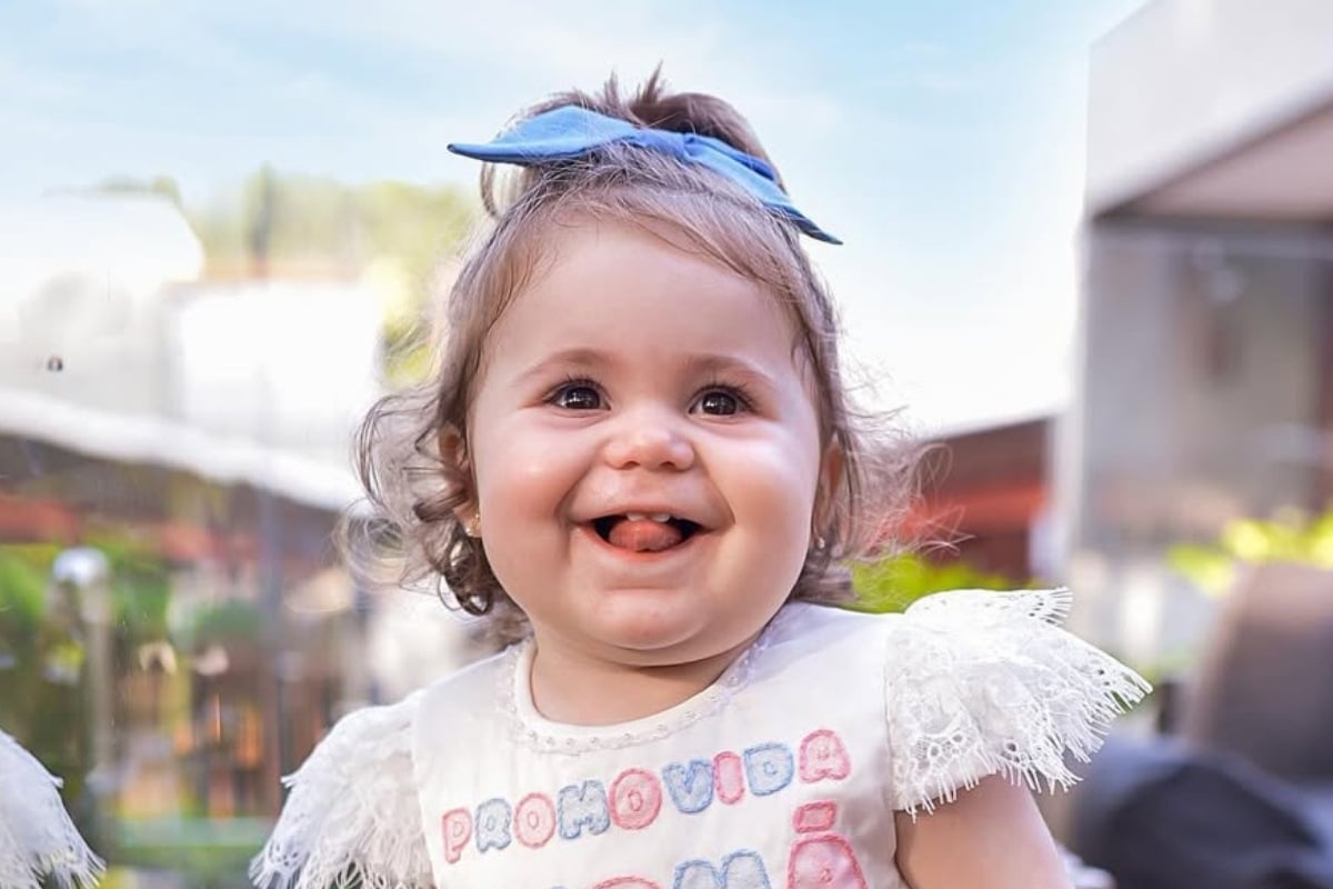 Uma adorável criança sorridente, com laço azul no cabelo, veste uma camisa com bordado colorido. Ao fundo, há um ambiente ao ar livre, desfocado. O rosto expressa alegria, capturando um momento de pura felicidade e ternura.