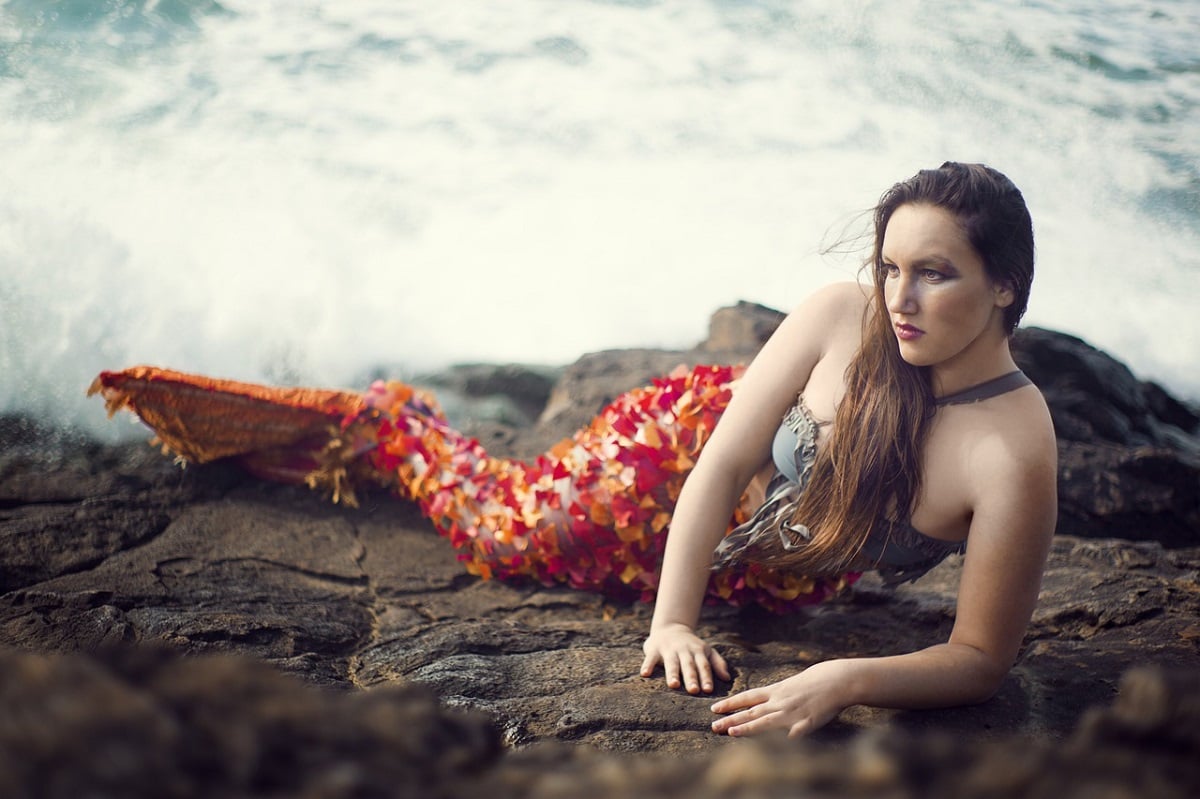 Foto de uma mulher com expressão séria deitada em rochas próximas ao mar. Ela está com fantasia de sereia e sua cauda é uma mistura das cores vermelho e laranja