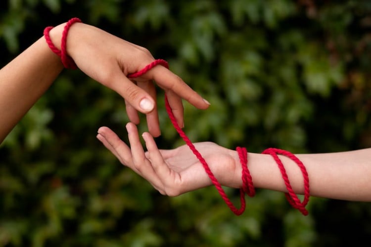 Duas mãos de pessoas de pele clara entrelaçadas por fio vermelho 