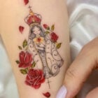 A tatuagem retrata uma figura estilizada de Nossa Senhora com uma coroa, cercada por rosas vermelhas e pequenos corações. O manto detalhado envolve a imagem, que exibe um coração no centro do peito. A composição é delicada e colorida, transmitindo um sentimento de paz e devoção. A mão ao lado destaca a suavidade do desenho.