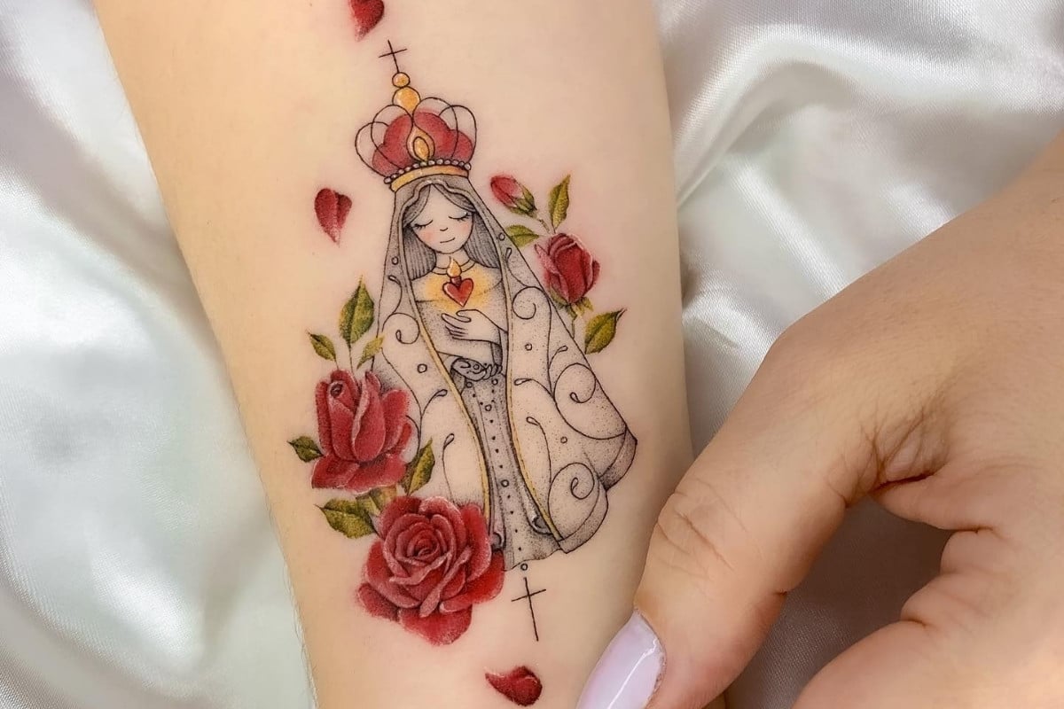 A tatuagem retrata uma figura estilizada de Nossa Senhora com uma coroa, cercada por rosas vermelhas e pequenos corações. O manto detalhado envolve a imagem, que exibe um coração no centro do peito. A composição é delicada e colorida, transmitindo um sentimento de paz e devoção. A mão ao lado destaca a suavidade do desenho.