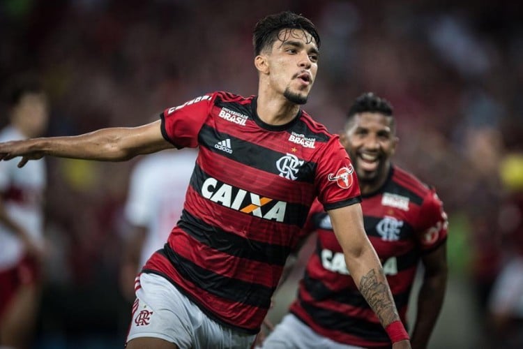 Lucas Paquetá com a camisa do Flamengo, de braços abertos em campo, com Rodinei desfocado ao fundo 