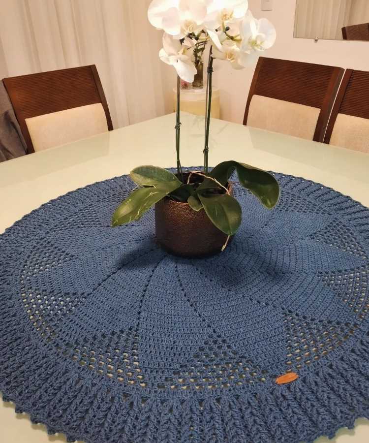 A imagem mostra uma mesa de vidro com um centro de mesa de crochê azul em formato circular. O crochê possui um padrão detalhado e bordas trabalhadas. No centro, há um vaso marrom com uma orquídea de flores brancas. Ao fundo, há cadeiras com encosto de madeira e estofado claro.