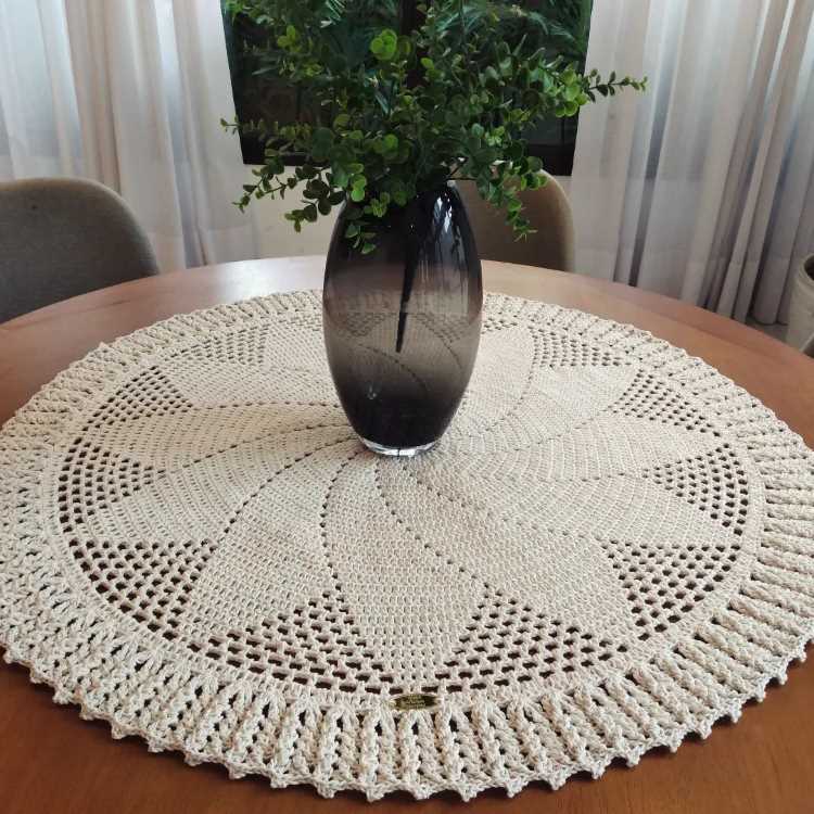 A imagem mostra uma mesa redonda de madeira com um centro de mesa de crochê em formato circular. O crochê é bege e possui um padrão detalhado com bordas trabalhadas. No centro, há um vaso de vidro escuro contendo um arranjo de folhas verdes. Ao fundo, cortinas claras e cadeiras estofadas são visíveis.