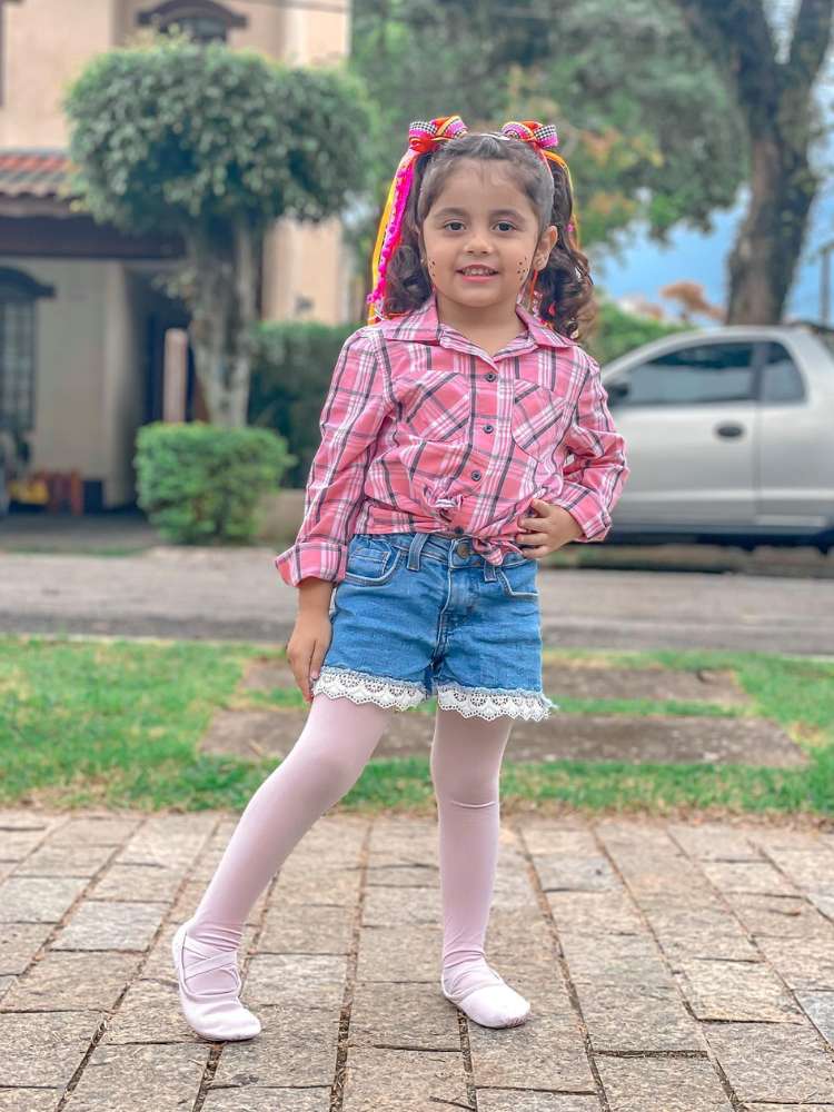 Menina de pele morena usando blusa xadrez rosa, shorts jeans, meia calça branca e fitas no cabelo