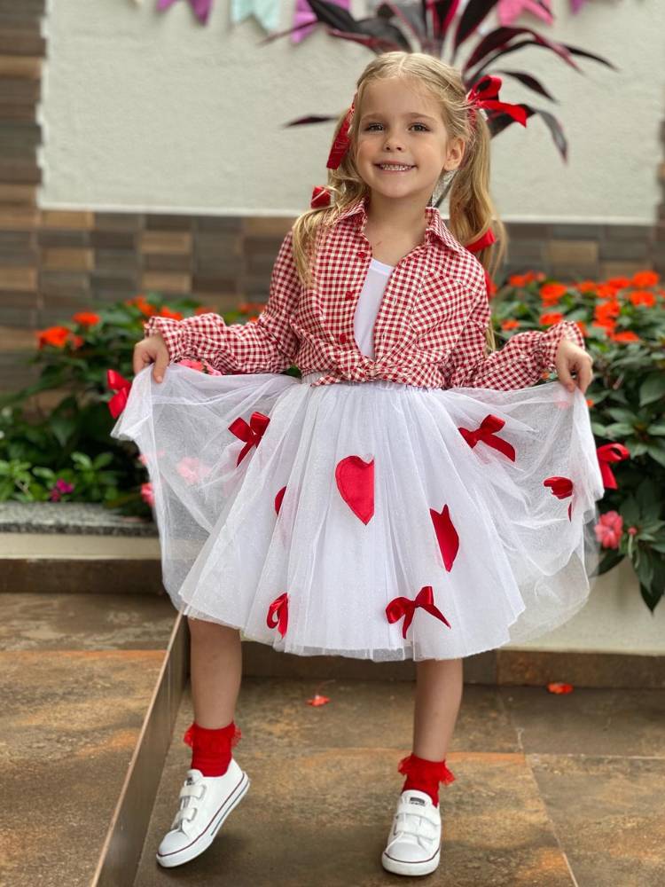 Menina de pele clara usando vestido branco com laços e corações e blusa xadrez branca e vermelha por cima