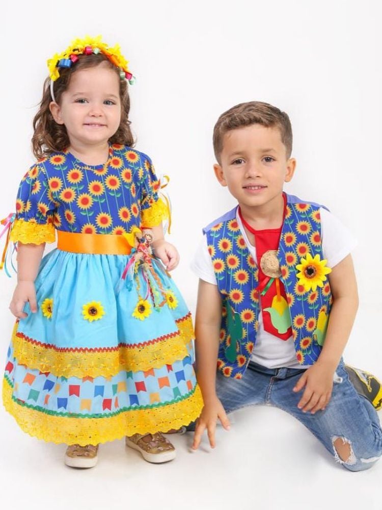 Menino e menina de pele clara com trajes juninos. Ela de vestido colorido com laços e girassóis e ele com colete azul de girassóis, blusa branca, gravata vermelha e calça jeans