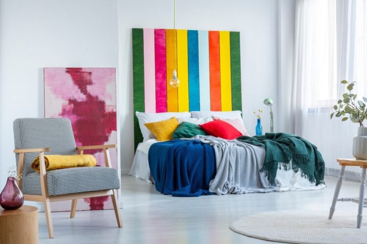 Foto de sala com quadro nas cores da bandeira LGBT, sofá branco com almofadas coloridas e cobertores, tapete bege, poltrona cinza e amarela e quadro rosa