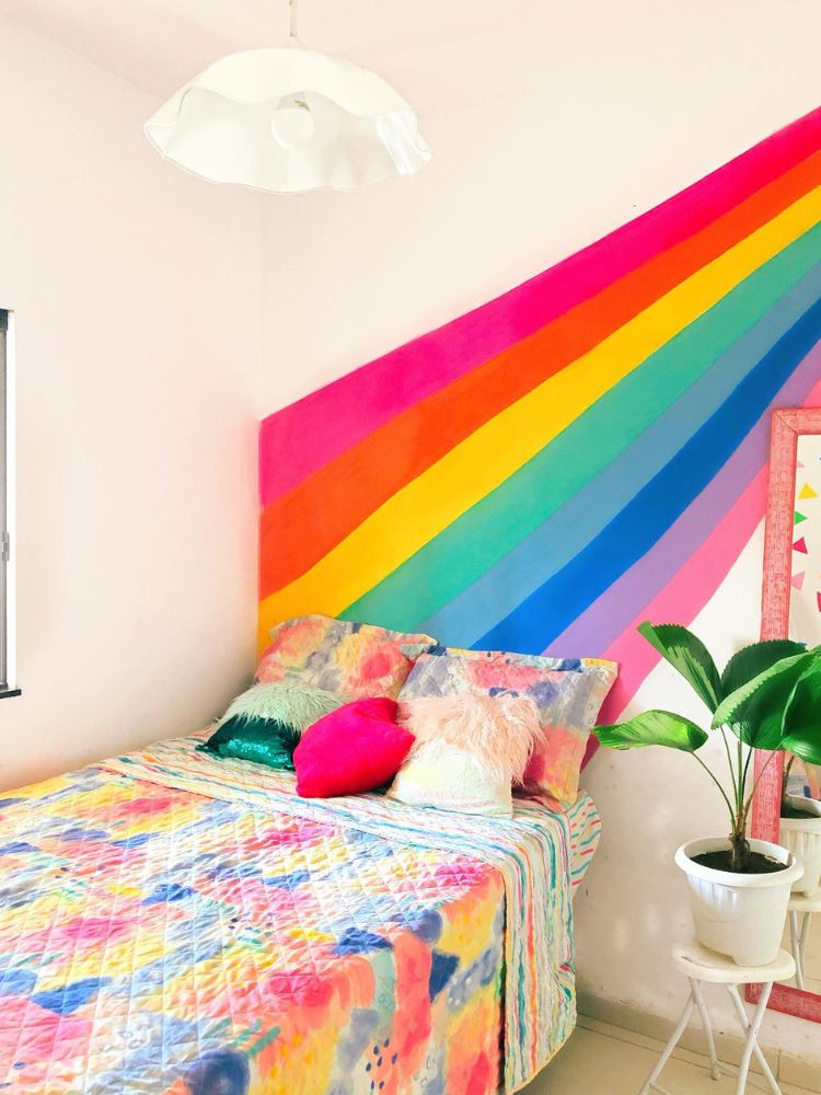 Quarto de parede majoritariamente branca, com pintura de cores do arco-íris e cama com lençol colorido