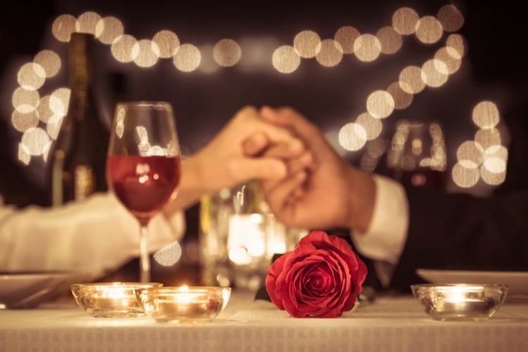 Duas pessoas de pele clara de mãos dadas sobre a mesa, com luzes, taça de vinho e flores 