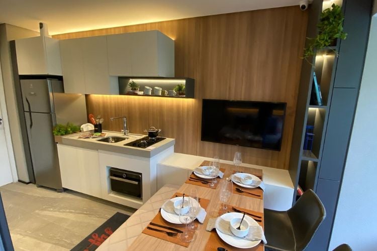 Apartamento pequeno decorado com painel de madeira que integra a sala de jantar com a cozinha, que possui armários claros. Ainda, há uma TV no painel e uma mesa de madeira 