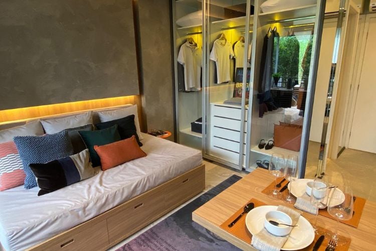 Apartamento com parede de cimento queimado, cama e sofá ao mesmo tempo, mesa de madeira com pratos e armário de roupas transparente