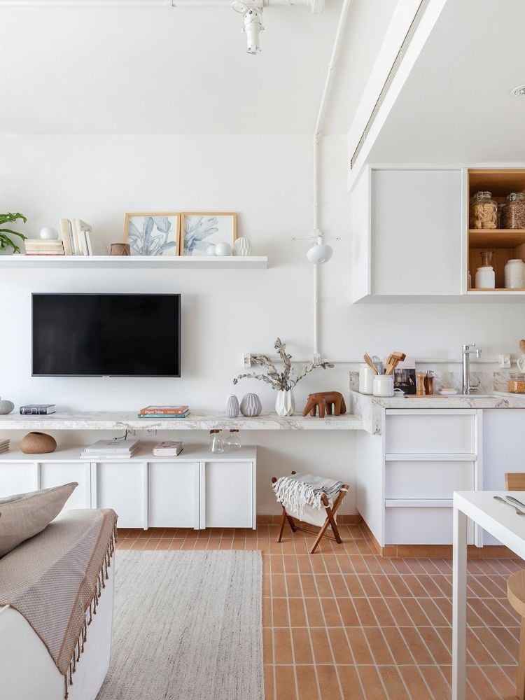 Apartamento com sala e cozinha integradas, em tons brancos e com o piso marrom. Os quadros e itens decorativos das prateleiras e móveis da sala são pequenos