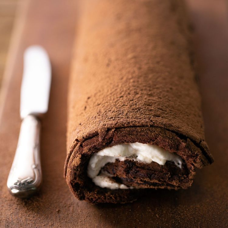 Rocambole de chocolate com creme branco em mesa de madeira ao lado de faca