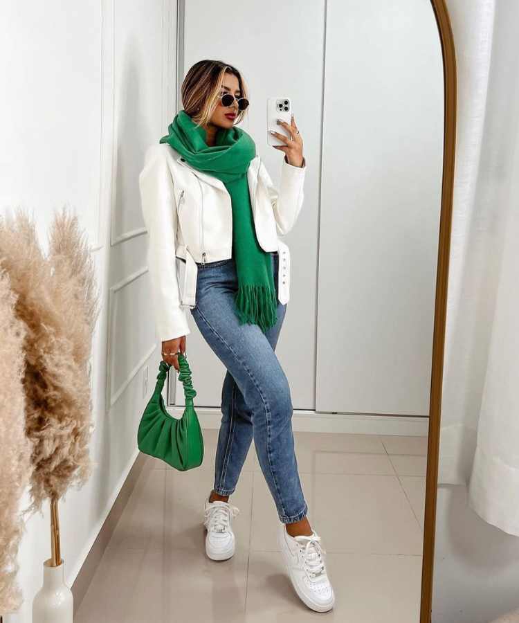 Imagem com fundo de quarto com espelho. No centro, mulher usando jaqueta de couro branca, cachecol verde, calça jeans, bolsa verde, tênica branco