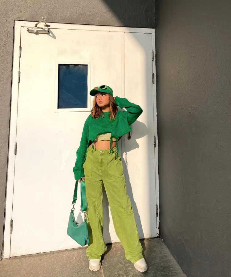 Imagem com fundo de parede e porta. No centro, mulher com look verde,  casaco, calça boné, bolsa e óculos na tonalidade, + bota e cropped bege