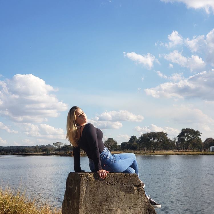 Foto da atriz de A Vida da Gente sentada em bloco de cimento grande de frente à lagoa