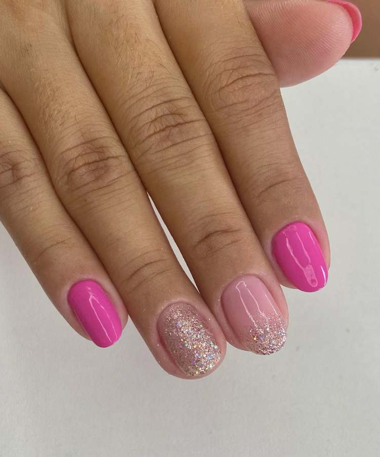 A imagem retrata um mão com unhas decoradas de rosa. Os esmaltes usados são de cor: rosa, nude com glitter prata e dourado. O esmalte está uniforme e brilhante. As unhas estão no formato redondo.