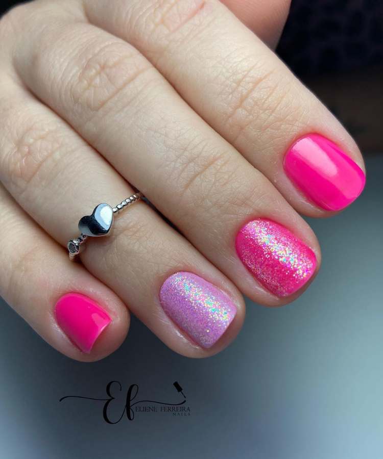 A imagem retrata um mão com unhas decoradas de rosa. Os esmaltes usados são de cor: pink, glitter e lilás. O esmalte está uniforme e brilhante. As unhas estão no formato redondo.