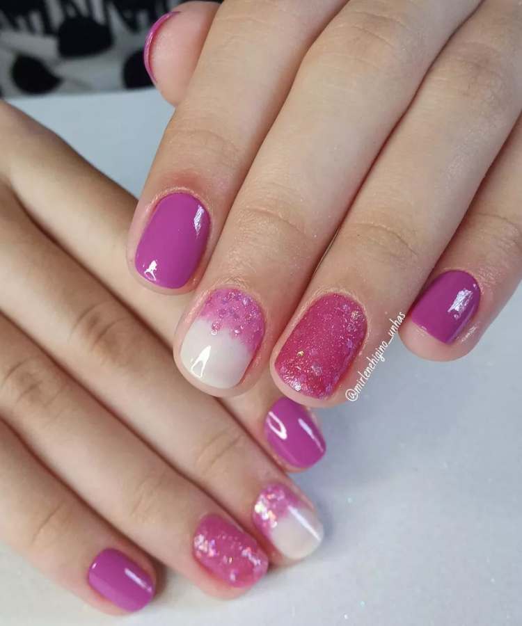 A imagem retrata um par de mãos com unhas pintadas de rosa. Os esmaltes usados são de cor: lilás, rosa, glitter rosa, branco. O esmalte está uniforme e brilhante. As unhas estão no formato redondo.
