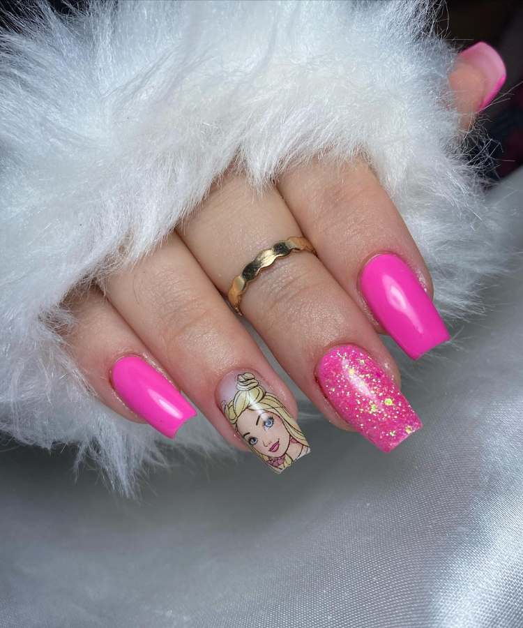 A imagem retrata um mão com unhas decoradas de rosa. Os esmaltes usados são de cor: pink, glitter dourado e rosa, adesivo de chiclete da Barbie. O esmalte está uniforme e brilhante. As unhas estão no formato quadrado.