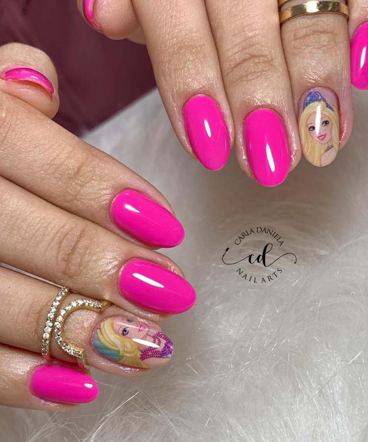 A imagem retrata um par de mãos com unhas pintadas de rosa - tendência Barbiecore. Os esmaltes usados são de cor: pink e nude com adesivo de chielete da Barbie. O esmalte está uniforme e brilhante. As unhas estão no formato redondo.