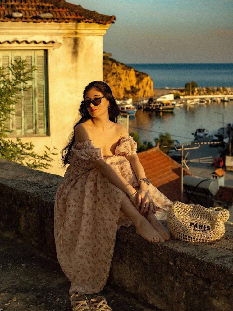 foto da estética tomato girl, com mulher de vestido florido sentada em mureta em cidade costeira da Europa