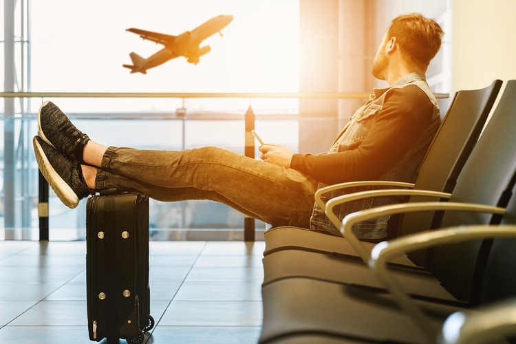 Homem sentado no saguão de um aeroporto com as pernas esticadas em cima de uma mala olhando um avião passar
