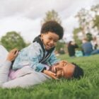 A imagem mostra uma criança repousando no colo de um adulto em um parque. Ambos estão sentados na grama, sugerindo um momento de conexão e relaxamento. A cena pode evocar o conceito de Dia dos Pais criativo, celebrando a relação entre pai e filho(a) ao ar livre.