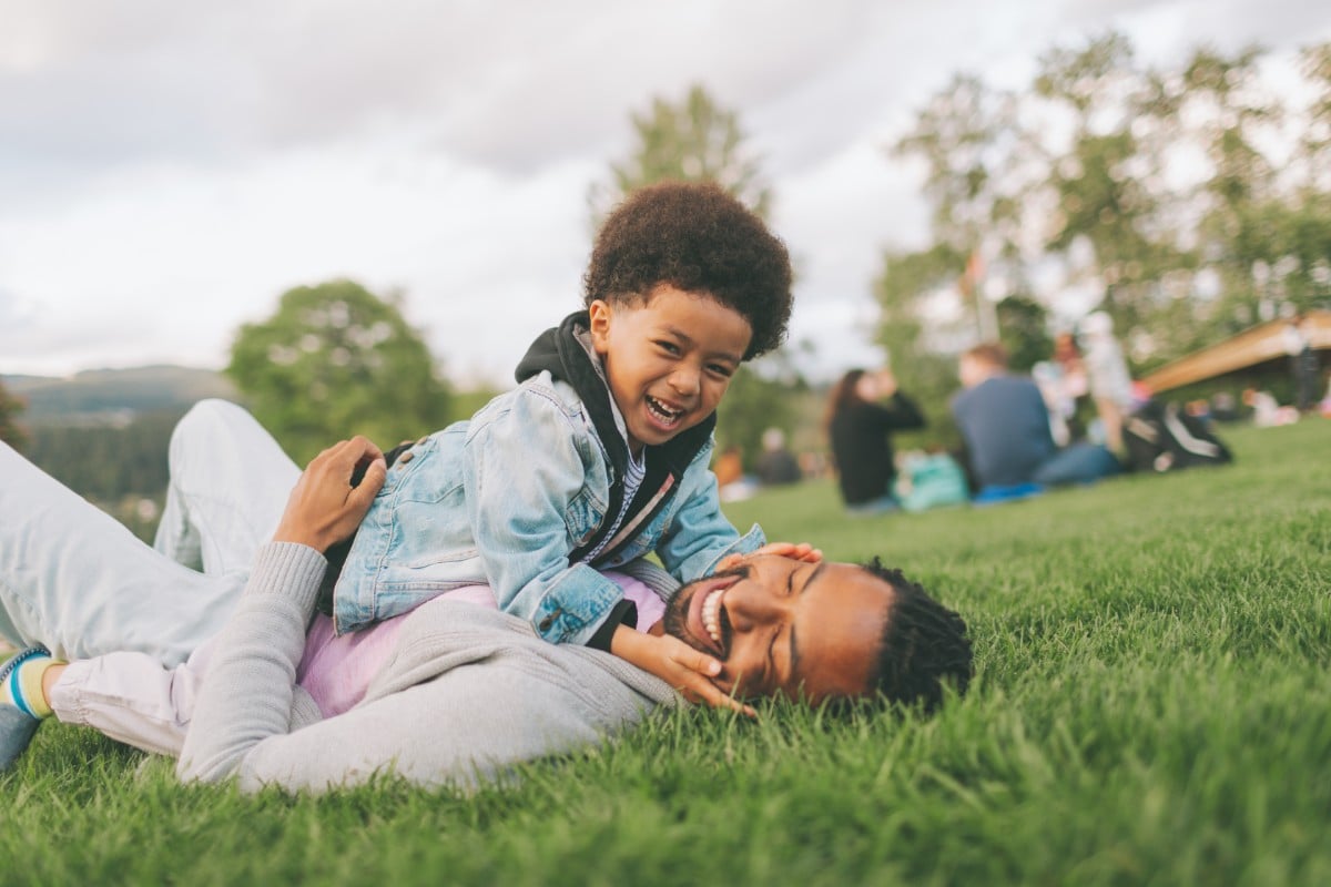 A imagem mostra uma criança repousando no colo de um adulto em um parque. Ambos estão sentados na grama, sugerindo um momento de conexão e relaxamento. A cena pode evocar o conceito de Dia dos Pais criativo, celebrando a relação entre pai e filho(a) ao ar livre.