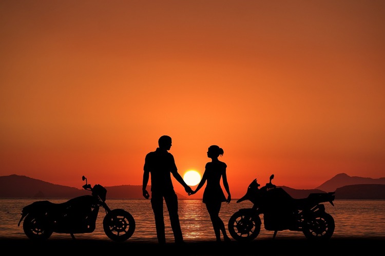 casal heterossexual de mãos dadas, ao lado de duas motos, em frente ao mar durante o pôr do sol