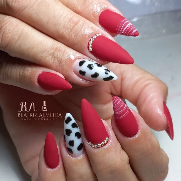 Mãos de pele clara com unhas stiletto médias, com esmalte vermelho fosco em todas exceto o anelar que possui animal print de vaca 