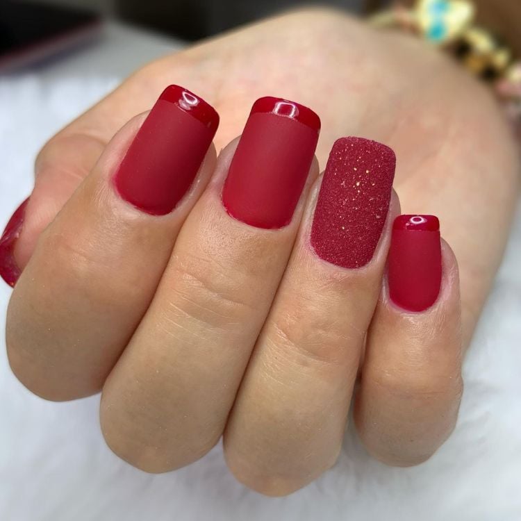 Mão de pele clara com unhas quadradas curtas com esmalte fosco, francesinha transparente brilhante e filha única vermelha com glitter 