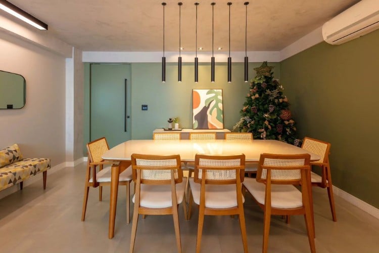 Sala de jantar com luminárias pendentes, parede verde musgo, mesa grande de jantar e árvore-de-natal ao fundo