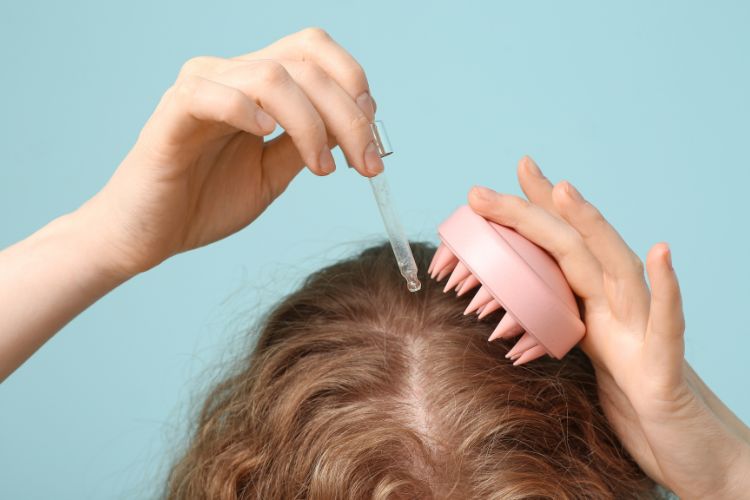 Mulher de pele clara aplicando sérum na raiz do cabelo e segurando massageador rosa