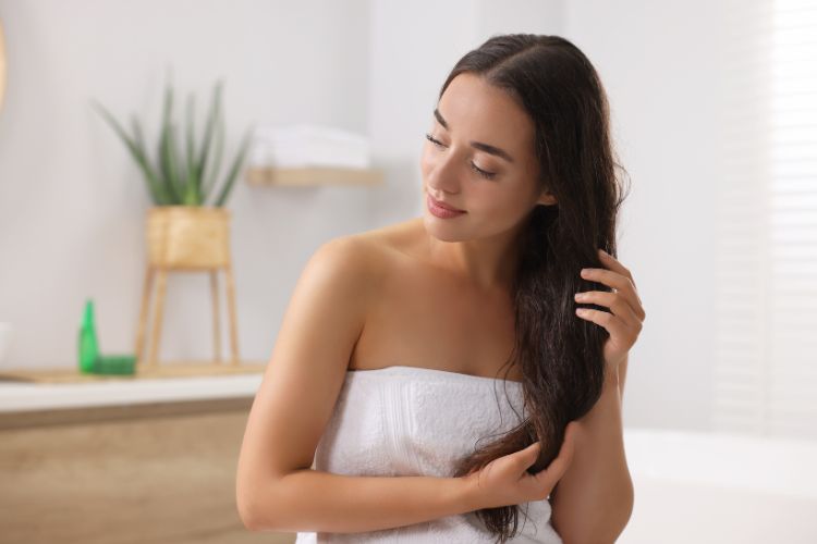 Mulher de pele clara em banheiro com plantas, amarrada em toalha branca e mexendo em cabelo longo