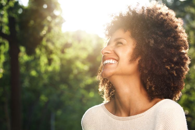 Mulher de pele negra com cabelo cacheado sorrindo ao sol e em meio a árvores