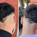 A imagem mostra dois cortes de cabelo masculinos com design moderno. Ambos têm cabelo curto nas laterais e mais cheio no topo. Cada corte apresenta linhas geométricas raspadas na parte inferior da cabeça, criando padrões estilizados e precisos. O fundo é uma parede de tijolos, e uma das pessoas usa máscara e brinco.