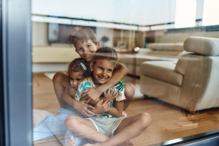 Foto de três irmãos abraçados em uma sala com sofá bege, se vendo no reflexo do vidro da porta