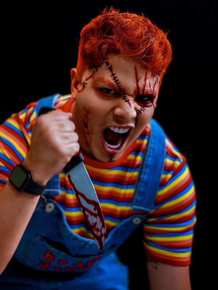 Homem de pele clara com blusa listrada colorida, macacão jeans, cabelo vermelho e maquiagem de Chucky, com machucados e costuras