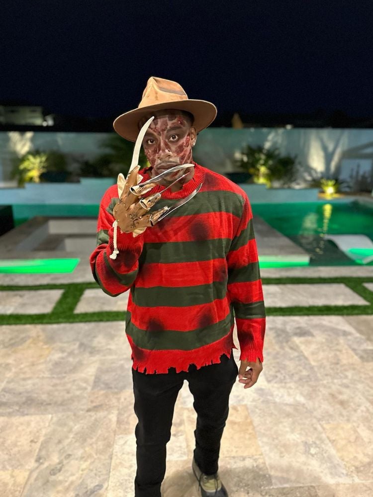 Homem de pele negra com chapéu bege, camiseta verde e vermelha listrada rasgada, calça preta, luva de Freddy Krueger com lâminas de mentira e maquiagem com cicatrizes 