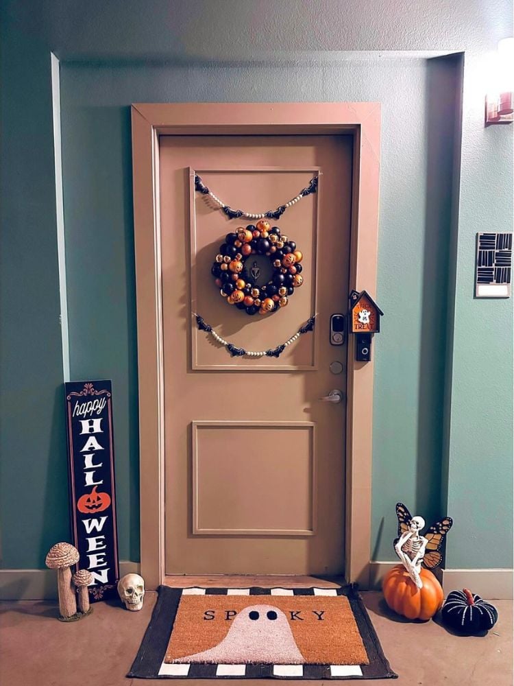 Entrada de apartamento com parede verde, porta marrom com guirlanda de Halloween, tapete de Halloween, abóboras, esqueletos e placa escrito "happy halloween"