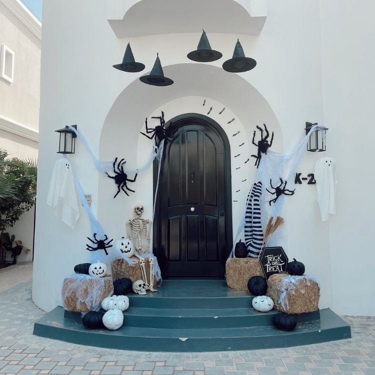 Entrada de casa com parede branca, porta marrom, escada azul e decorações em preto e branco, como abóboras, chapéus de bruxa, canelas de bruxa e mais 