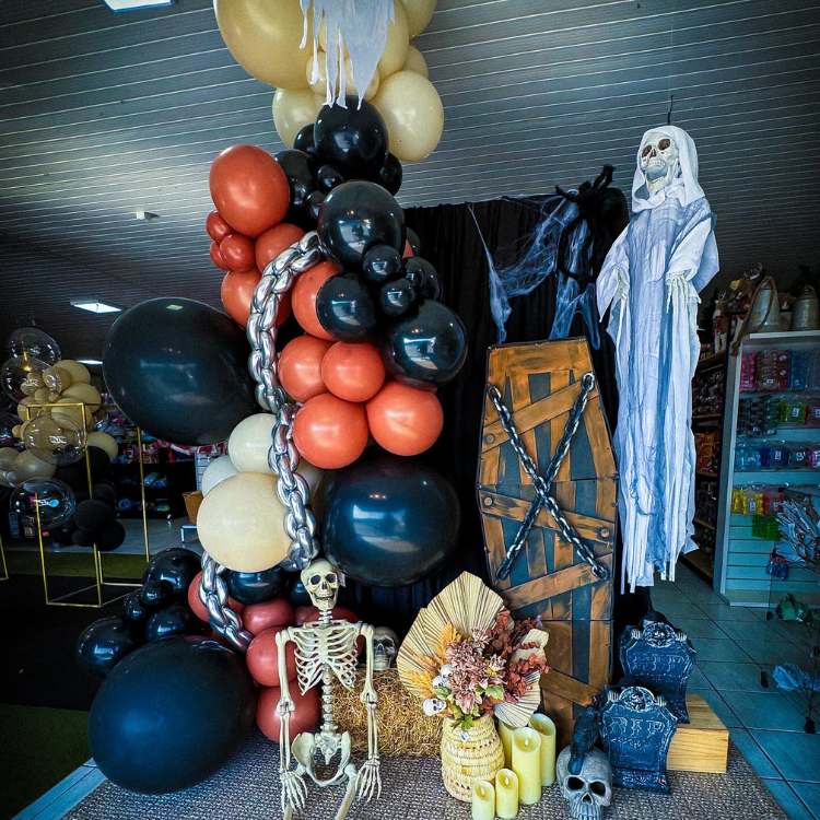 Vitrine de Halloween com tema de cemitério, com caixão, lápide, fantasma, esqueleto e bexigas laranajs e pretas 