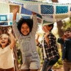 A imagem mostra quatro crianças brincando ao ar livre, segurando uma colcha colorida acima de suas cabeças. Elas estão sorrindo e parecem estar se divertindo muito. O ambiente é cercado por árvores, sugerindo que estão em um parque ou bosque. A luz do sol filtra pelas folhas, criando uma atmosfera alegre e vibrante. As crianças estão vestidas de forma casual e a cena transmite uma sensação de felicidade e liberdade.