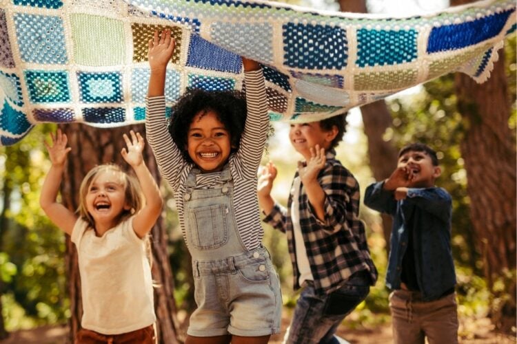 A imagem mostra quatro crianças brincando ao ar livre, segurando uma colcha colorida acima de suas cabeças. Elas estão sorrindo e parecem estar se divertindo muito. O ambiente é cercado por árvores, sugerindo que estão em um parque ou bosque. A luz do sol filtra pelas folhas, criando uma atmosfera alegre e vibrante. As crianças estão vestidas de forma casual e a cena transmite uma sensação de felicidade e liberdade.