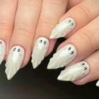 A imagem mostra unhas decoradas com desenhos de fantasmas. As unhas são pontiagudas e pintadas de branco, com detalhes de olhos pretos e bochechas rosadas, criando um efeito lúdico e divertido. O design é detalhado, lembrando pequenos fantasmas estilizados, ideal para uma temática de Halloween ou para quem busca um visual criativo e único.