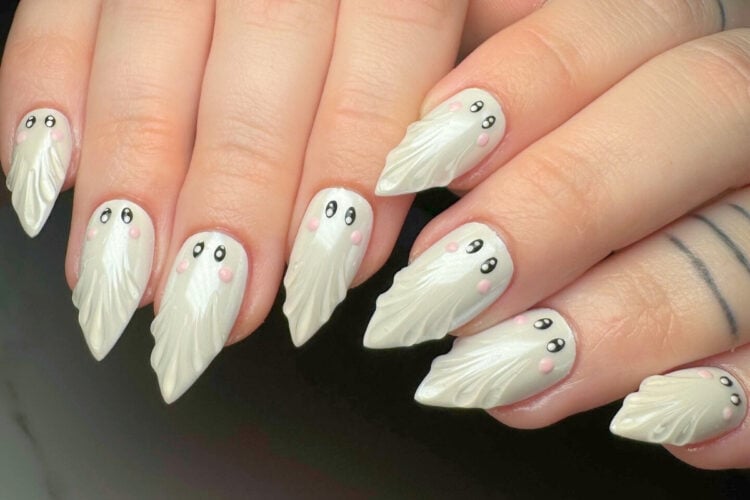 A imagem mostra unhas decoradas com desenhos de fantasmas. As unhas são pontiagudas e pintadas de branco, com detalhes de olhos pretos e bochechas rosadas, criando um efeito lúdico e divertido. O design é detalhado, lembrando pequenos fantasmas estilizados, ideal para uma temática de Halloween ou para quem busca um visual criativo e único.