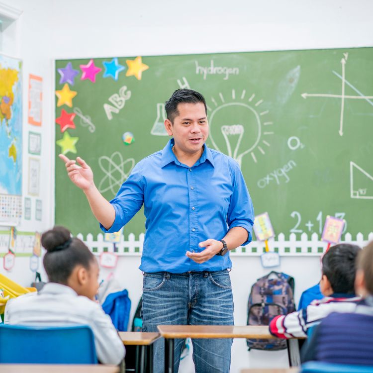 Professor asiático usando camisa azul e calça jeans e dando aula, com lousa preenchida ao fundo 